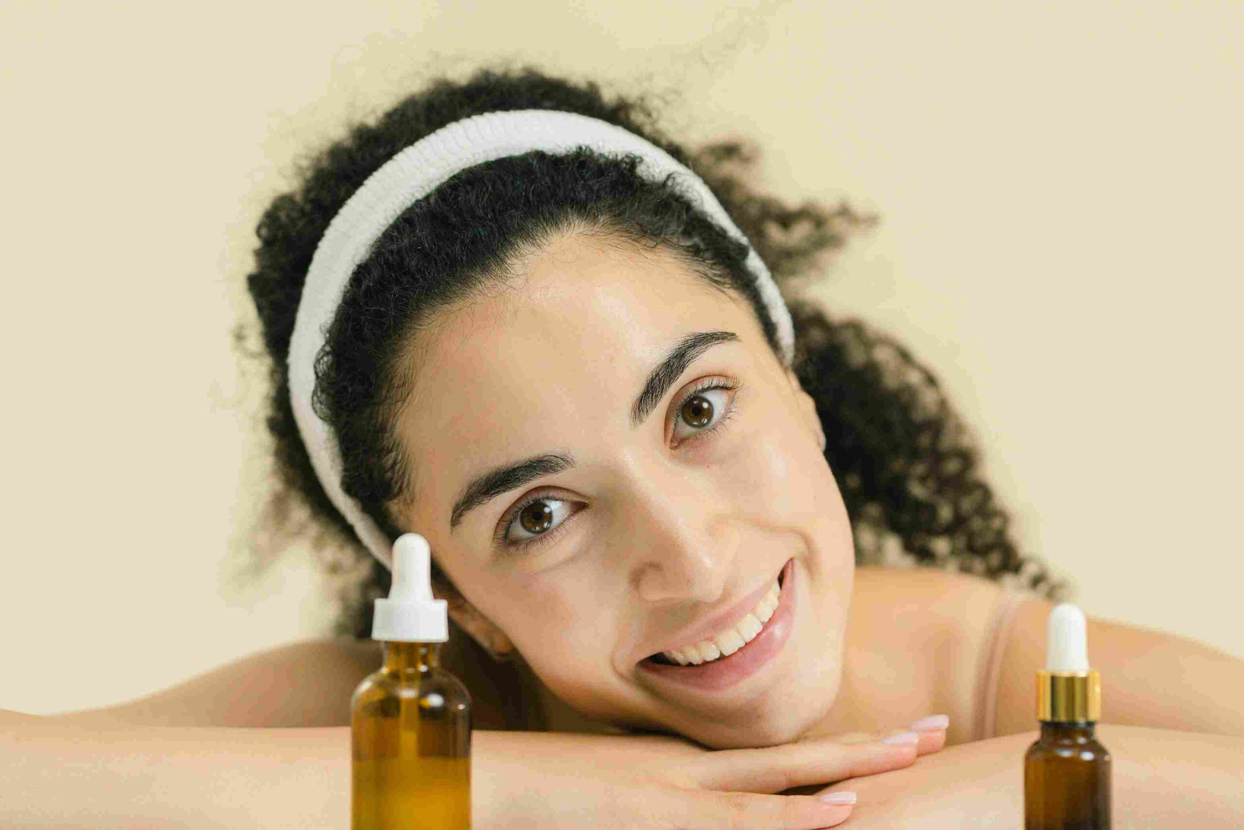 A smiling woman with curly hair, wearing a white headband, resting her face on her hands with two bottles of face serum in front of her. How to Use Face Serum for Dry Skin effectively.