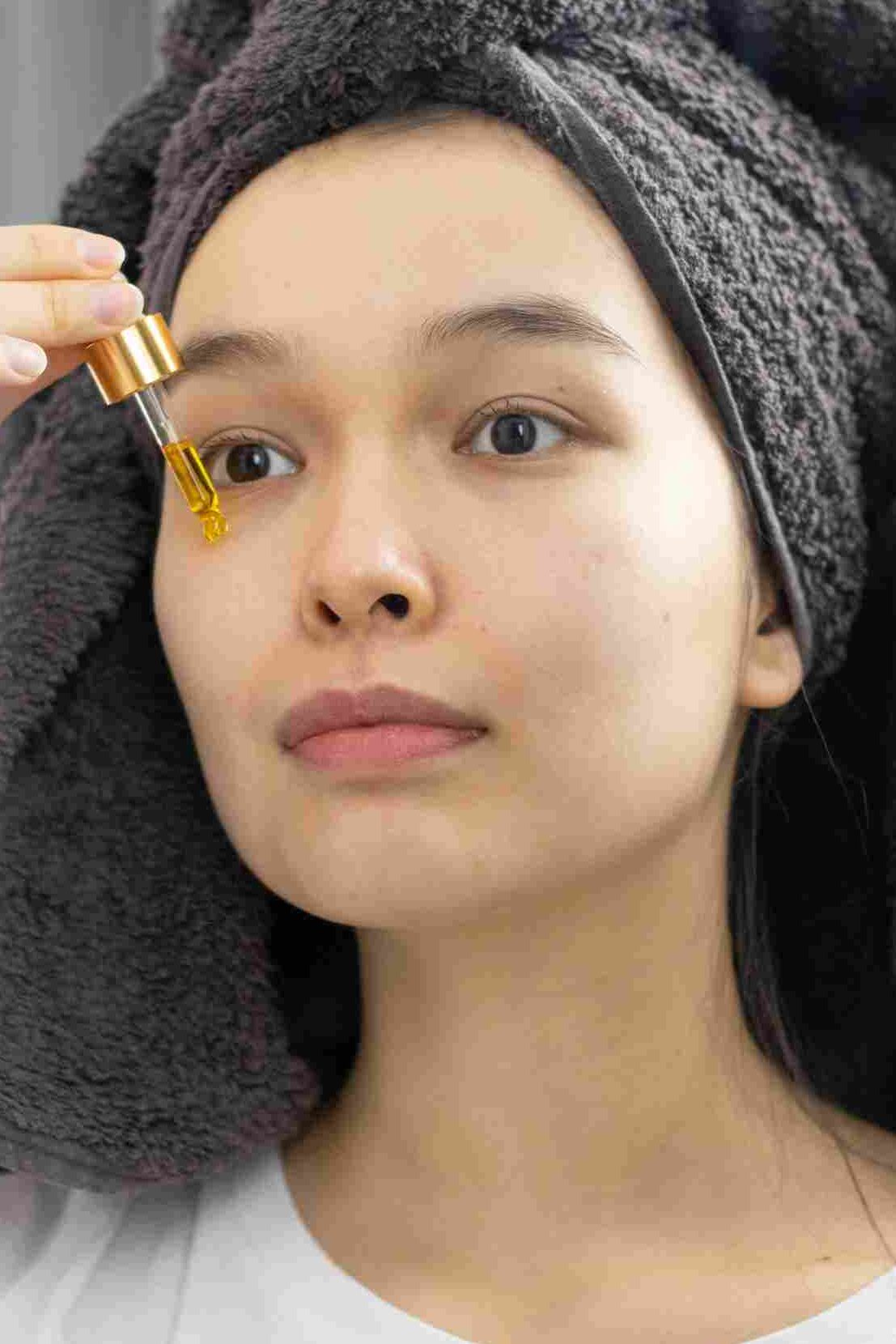 A woman with a towel wrapped around her head, applying face serum with a dropper while looking into a mirror. How to Use Face Serum for Dry Skin correctly. | womenluxlife.com