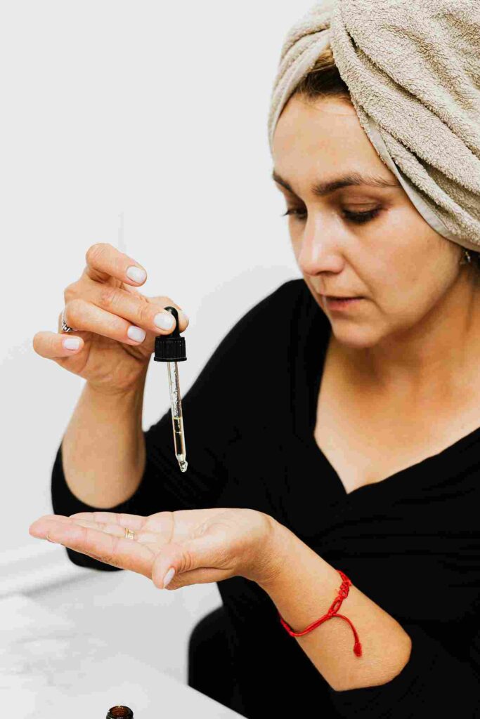 A woman with a towel wrapped around her head is applying serum to her palm using a dropper. She is wearing a black top and a red string bracelet. The image represents skincare application, specifically highlighting the topic "Serum vs. Cream for Dry Skin. | womenluxlife.com